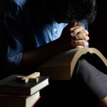 Osoba u molitvi nad Biblijom, s knjigama i križem u prvom planu, u tihoj i intimnoj atmosferi.