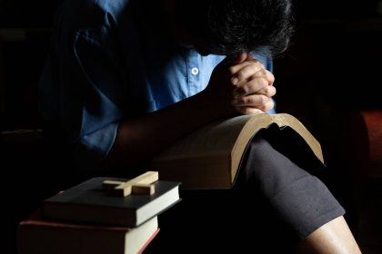 Osoba u molitvi nad Biblijom, s knjigama i križem u prvom planu, u tihoj i intimnoj atmosferi.