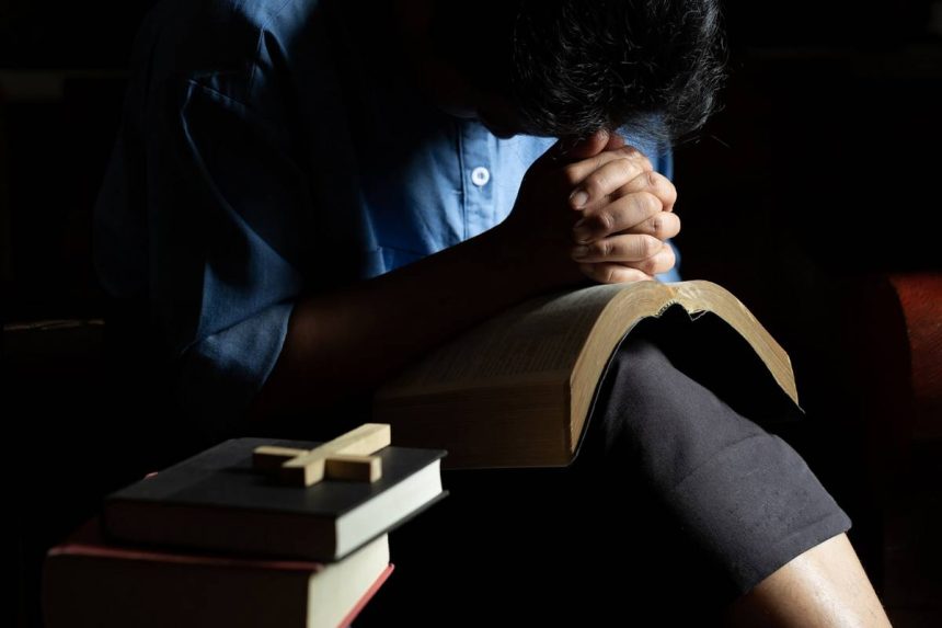 Osoba u molitvi nad Biblijom, s knjigama i križem u prvom planu, u tihoj i intimnoj atmosferi.