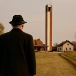 Čovjek, okrenut leđima, u crnom kaputi i sa crnim šeširom na glavi gleda u pravcu lokalne crkve u ravnici.