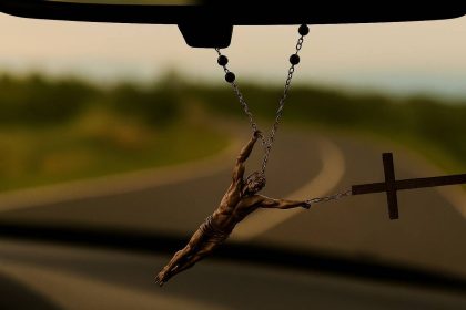 Krunica visi s retrovizora automobila, nagnuta zbog zavoja u vožnji. Isus, prikazan kao stvaran ljudski lik, otkinut je s križa i objeručke se drži za krunicu iznad križa, kao da ga inercija pokušava odbaciti. U pozadini se kroz vjetrobransko staklo vidi zamućena zavojita cesta.