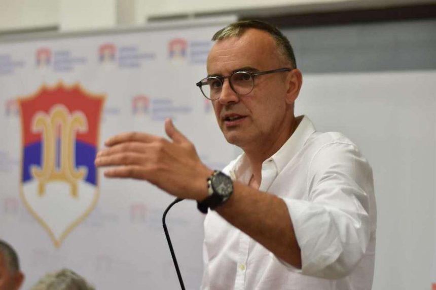 Političar Babalj za govornicom u bijeloj košulji s podignutom rukom na kojoj je sat