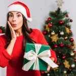 Žena u crvenom drži zeleni paket. U pozadini okićen novogodišnji bor.