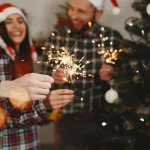 Muškarac i žena u novogodišnjem raspoloženju pored okićenog bora u ruci drže prskalice.