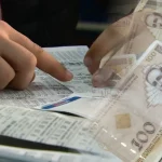 Dvije ruke iznad kladioničarskog tiketa popunjavaju tiket. Desno konvertibilne marke.