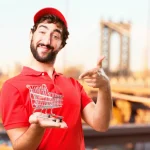 Trgovac u crvenoj majici s osmjehom na licu drži u ruci minijaturna trgovačka kolica.