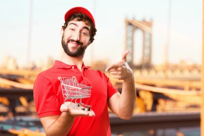 Trgovac u crvenoj majici s osmjehom na licu drži u ruci minijaturna trgovačka kolica.