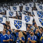 Navijači Bosne i Hercegovine u plavim dresovima mašu zastavama s ljiljanima na prepunim tribinama stadiona.